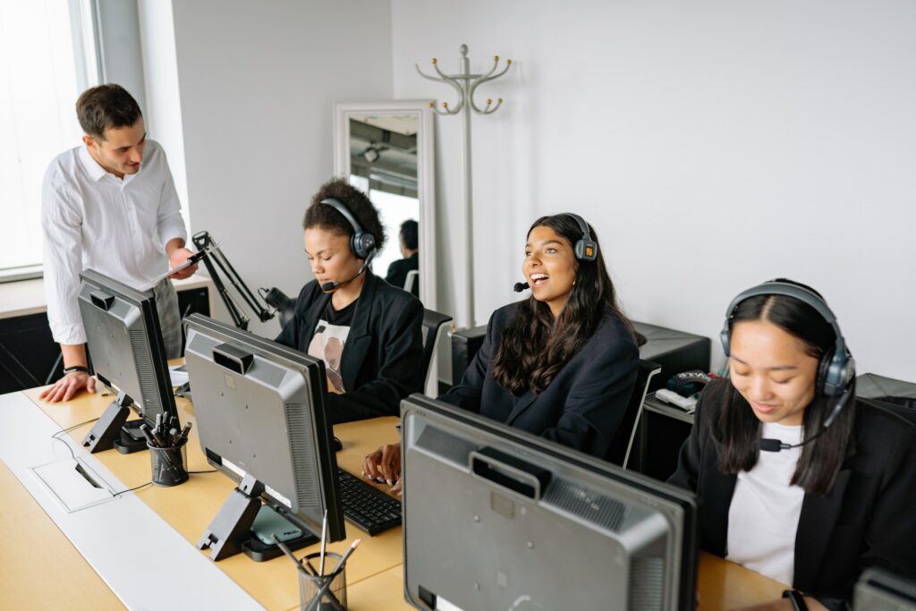Drie vrouwen zitten elk achter een computer met headsets op en praten met een man die naast het bureau staat met een tablet z'n hand.